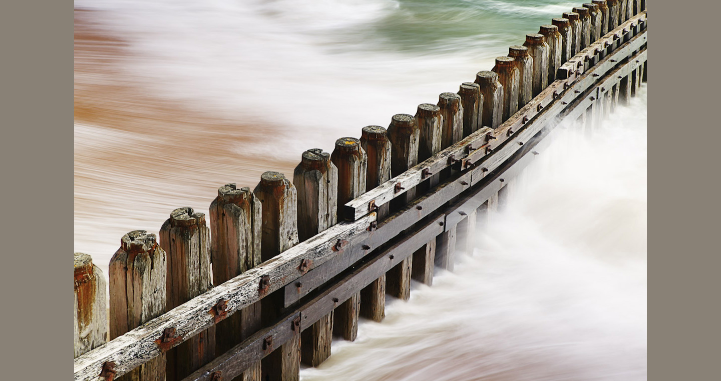 A snaking timber groyne contrasts with golden sand, white waves and green sea
