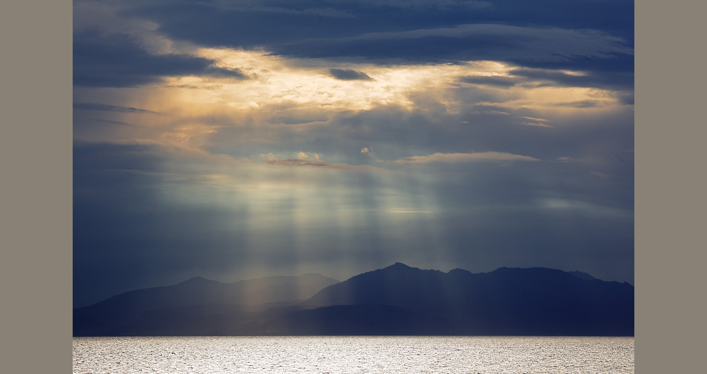 Sunlit break in grey clouds silhouettes a dark Arran behind sparkling sea